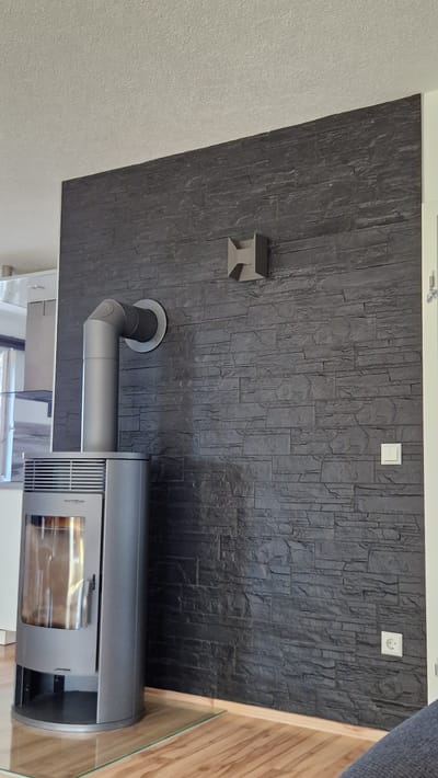 The image shows a modern living room scene in Ichenhausen, Germany. A contemporary grey wood-burning stove is positioned in the lower left corner, with a metallic flue pipe extending upwards and connecting to the wall. The wall behind the stove is covered in dark grey, textured stone veneer. Mounted on the stone wall, slightly to the right of the flue pipe, is a decorative, geometric, metallic object. Further to the right on the wall are a light switch and an electrical outlet. The floor is made of light-colored wooden planks, and a clear glass plate is placed in front of the stove for protection. The ceiling is a light color with a textured finish. The overall impression is one of a well-designed, modern interior.