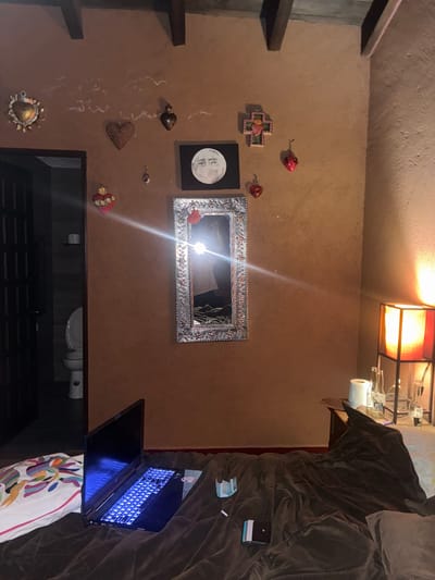 The image shows a room in what appears to be a home. On the left wall are wooden shelves and various decorative items, including a cross, moon art, hearts, and a sun. A framed mirror hangs on the wall. Below the wall decorations is a dark surface that appears to be the top of a cabinet or console. A toilet and sink are positioned below the mirror and cabinet. A laptop is open and illuminated on the right side of the image, resting on bedding and pillows. A dark blanket covers the bed. The room is lit by a square lamp on the ceiling. The specific location in the city of Cañada de Madero, Mexico, cannot be determined based solely on this image. No people are visible.