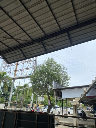 This image captures a scene under a corrugated metal roof, looking upwards and outwards towards an outdoor area. The foreground is dominated by the underside of the roof, supported by dark metal beams. Beyond the roof, a treed area with lush green foliage is visible, with a bright, overcast sky. In the middle ground, a complex metal structure, possibly an antenna or scaffolding, rises above the trees.

Further back, the scene transitions to a more urban or market-like setting. A white building with multiple levels and windows is visible, with a prominent sign that reads "SINAR PONSEL". Several cars and a motorcycle are parked in front of this building. There are also people walking around, suggesting a busy public space. Some palm trees and other greenery are interspersed with the buildings.

The lighting suggests it is daytime, likely overcast given the diffused light. The presence of people, vehicles, and market stalls indicates an active commercial or community area. The overall impression is one of a functional, possibly bustling, outdoor environment in a tropical or subtropical region, as hinted by the palm trees and the general atmosphere. The image is oriented vertically, giving a sense of looking up and out.