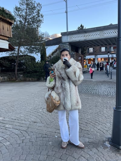 The image displays a woman standing on a cobblestone street in Gstaad, Switzerland, during daylight. She is centrally positioned, wearing a light-colored fur coat, white wide-leg pants, and light brown loafer-style shoes. Black gloves cover her hands; her right hand holds a white disposable cup to her mouth, while her left hand holds a smartphone featuring a blue PopSocket. A light brown, fur-trimmed bucket bag with white embroidery hangs from her left shoulder. Dark sunglasses rest on top of her head.

To her left, a wooden chalet-style building with a distinctive roof and evergreen trees are visible. Further back on the left, a man in a dark jacket holds a phone. In the mid-ground, an ornate concrete or stone overpass, decorated with repeating circular motifs, spans the scene, with overhead power lines crossing above it. Beyond the overpass to the right, a commercial street features multiple pedestrians, traditional buildings, and a clearly visible "Hermes" sign. Snow-capped mountains are present in the distant background under a clear blue sky. The ground is paved with grey cobblestones arranged in an arc pattern. A black lamppost is partially visible on the far right side of the frame.