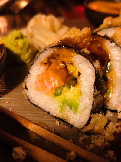 A close-up, high-angle shot captures a close-up of sushi rolls served on a wooden surface. The sushi is breaded and fried, with visible pieces of salmon and avocado inside. A dark sauce, likely teriyaki or eel sauce, is drizzled over the rolls. Two wooden chopsticks rest diagonally across the bottom of the frame. In the background, out of focus, is a small mound of green wasabi and what appears to be a small dish of dipping sauce. The lighting is warm and dim, suggesting an indoor setting, possibly a restaurant. There are no people visible in the image. The image does not contain any information that would indicate its location in Be'er Sheva, Israel.