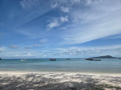 This is a photograph of a tranquil beach scene in Koh Toch Village, Cambodia. The foreground is dominated by a white sandy beach, partially shaded by dappled sunlight filtering through unseen trees. In the mid-ground, the calm, clear blue-green waters of the sea stretch towards the horizon. Several boats are visible on the water. Closest to the shore, a white speed boat is moving from left to right. Further out, there are two traditional fishing boats, one blue with a red canopy and passengers visible, and a yellow boat. To the right, another green boat is moored, with a forested island visible in the far distance. The sky is a bright blue, dotted with scattered white cumulus clouds and wispy cirrus clouds. The overall impression is one of peace and natural beauty on a sunny day.