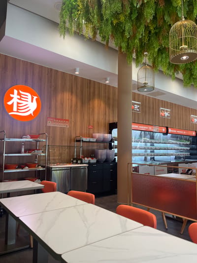 The image depicts the interior of a restaurant in Bottrop, Germany. The scene is a well-lit dining area with a modern aesthetic. In the foreground, several white marble-patterned tables are set, with bright orange chairs visible around them. The walls are paneled with wood, and artificial green plants hang from the ceiling, adding a touch of nature.
Several elements suggest a self-service or fast-casual dining experience. There are shelves stocked with red bowls, stacks of plates, and what appear to be ingredient containers or serving dishes. A refrigerated display case runs along the back wall, filled with pre-portioned meals or ingredients in clear containers, suggesting a food assembly line. Signage in Chinese and English indicates "PICK-UP AREA" and "DISHES PREPARING." There are also decorative birdcages hanging from the ceiling, one containing a light fixture. A cylindrical pillar stands in the middle of the space. The overall atmosphere is clean and organized. There are no visible people in the image.