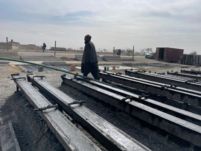 This image depicts a construction site or precast concrete manufacturing yard in Nakhonay, Afghanistan, under an overcast sky. In the foreground, multiple long, rectangular metal molds are arranged in parallel rows on mounds of dirt and aggregate. These molds contain freshly poured, wet concrete, with some showing visible rebar or anchoring points. A green hose runs horizontally across several of these molds.

In the midground, an adult male, with a beard and wearing a traditional dark tunic, loose trousers, and a cap, walks from left to right, looking towards the right side of the frame. He is positioned behind the concrete molds.

The background reveals a dusty, open environment. To the left, a person on a bicycle travels along a dirt path or road. Further back, low-rise, unpainted structures and a sparse fence line are visible. To the right, a light-colored sedan is parked next to a large, red metal shipping container that appears to be positioned on its side or tilted. Stacks of what appear to be finished concrete panels or blocks are visible near the container. The distant horizon features sparse, bare trees beneath a hazy sky.
