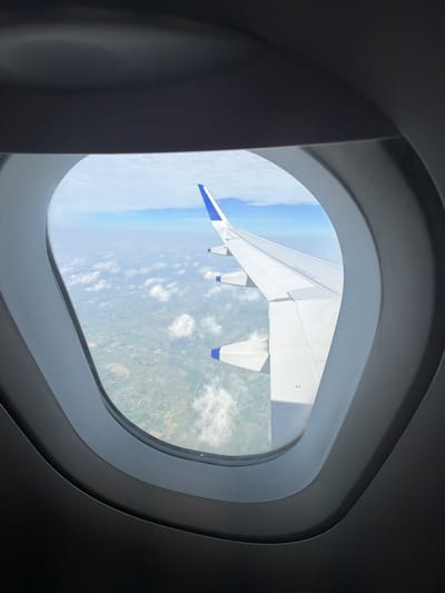 A view from an airplane window shows the wing of the aircraft extending to the right. Below the wing, a patchwork of green fields and scattered buildings is visible, indicative of the landscape surrounding Mohan, India. The sky is a bright blue, dotted with scattered white clouds. The interior of the airplane cabin is dark and frames the window view.