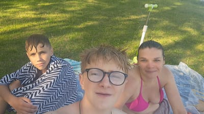 This is a selfie taken outdoors in a grassy area, likely a park or backyard, in Parchim, Germany. Three individuals are present. On the left, a young male is wrapped in a black and white striped towel, looking towards the camera with a neutral expression. In the center, a slightly older male with blond, spiky hair and glasses is in the foreground, also looking at the camera. On the right, a woman wearing a pink bikini smiles at the camera. Behind the individuals, a scooter with a green handlebar is propped upright on the grass. The ground is covered in green grass, and sunlight casts shadows across the scene. There are also towels and a plastic container visible on the ground.