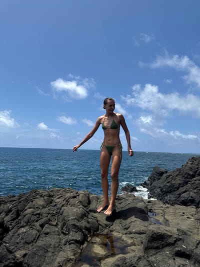 The image shows a woman in a green bikini standing on a rocky cliff. The sky is blue with scattered white clouds, and the ocean is visible behind her. The rocks appear to be volcanic and dark in color. The location is not identifiable as Santa Monica, Philippines based on the visual content.