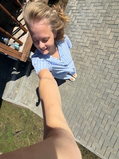 The image, taken from a high angle, shows a woman in a blue and white striped shirt standing on a brick-paved area with a grassy section visible on the right. She is wearing patterned slip-on sandals. Part of a wooden railing with a water bottle on it is visible in the lower left corner. The photograph appears to be a selfie, as the arm holding the camera is in the foreground. The location is likely outdoors and in a residential area, but specific details to confirm it is Kamennoye, Russia, are not present in the image.