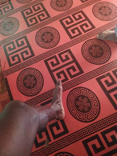 The image displays a close-up, elevated view of a human leg and foot resting on a red surface with repeating black geometric patterns, located in Unguwan Rimi, Nigeria. The surface is patterned with large black square Greek key designs and circular Greek key designs, separated by horizontal black Greek key borders. A bare human leg, exhibiting dark skin tone, is visible from the knee down, positioned in the lower-left to mid-left section of the frame, with the foot extended towards the upper center. A partial view of a second bare foot, appearing lighter in skin tone, is visible in the upper-right corner of the frame. Small dark specks are present on the red patterned surface.