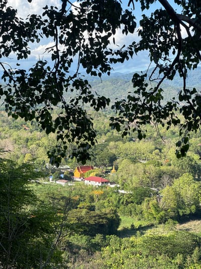 The image, taken in Ban Sahakon 2, Thailand, shows a lush green mountainous landscape. In the foreground, the silhouetted branches and leaves of a tree obscure part of the view. A collection of buildings, possibly a temple complex, with red and gold roofs, is situated in the middle ground. The surrounding vegetation consists of dense green trees. A blue sky and distant mountains can be seen through gaps in the tree branches.