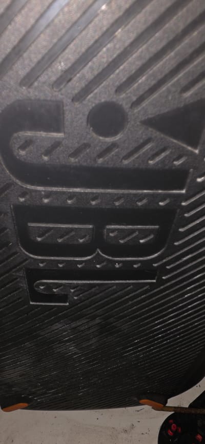 The image shows the sole of a black shoe, featuring a patterned tread. In the center of the sole, there is a raised design that appears to be the letters "BB" within an ornate border. The shoe sole is lying on a light-colored, dusty surface, possibly concrete. The bottom edge of the image shows a small orange protrusion from the shoe and a dark object with red circular details, potentially part of a wheel or a toy. The image does not contain any identifiable landmarks or elements that would suggest a specific location within the city of Hadera, Israel.