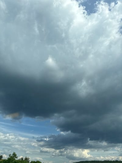 This is a photograph of a stormy sky over a landscape. The majority of the image is filled with dark, heavy clouds, suggesting an impending storm. There are breaks in the clouds revealing patches of blue sky. The bottom portion of the image shows a tree line with some greenery, hinting at land, possibly hills or a forest. There is no visible evidence of people or specific structures to indicate the location is the city of Rajana, Bulgaria. The lighting is dim and overcast due to the cloud cover.