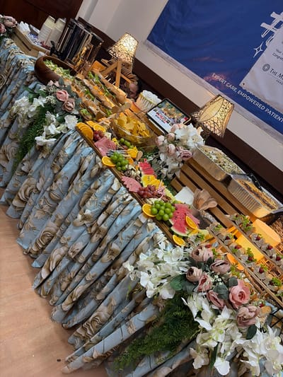 An indoor catering setup features a long table draped with a blue floral tablecloth. The table is laden with various food items, including sandwiches on skewers, fruit platters with watermelon, oranges, grapes, and berries, a salad in a wooden bowl, bowls of chips, and two foil catering pans containing pasta dishes. Decorative elements include artificial flowers, greenery, and two ornate lamps. In the background, a large blue banner with white text is visible, and a coffee urn and stacks of paper cups are placed on a wooden crate. A person's face is partially visible behind the table. The scene appears to be set up for an event or gathering. The presence of the "The M... Pang..." text on the banner, likely an acronym and a place name, combined with the general aesthetic, suggests the location could be within a venue in Dagupan City, Philippines, possibly related to a corporate or organizational event.