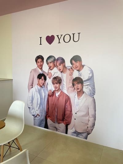 The image displays an indoor commercial space, presumed to be in Anápolis, Brazil, featuring a large wall graphic. The graphic, covering a substantial white wall, depicts a group portrait of seven male individuals identified as the K-pop group BTS, arranged in a multi-level pose. Above the figures, the phrase "I ❤️ YOU" is printed in black capital letters, with a purple heart symbol replacing "love." The individuals are dressed in coordinated light-colored attire, including suits, jackets, and pants, in shades of white, cream, light pink, and salmon. Several figures are smiling, and some are shown with arms around others. The floor consists of light-colored tiles. In the foreground on the left, a white, modern-design chair with wooden legs is visible, positioned next to a small, round table with a wooden top. A portion of a light yellow wall is also present on the far left. The precise location or address within Anápolis cannot be determined from the visual evidence.