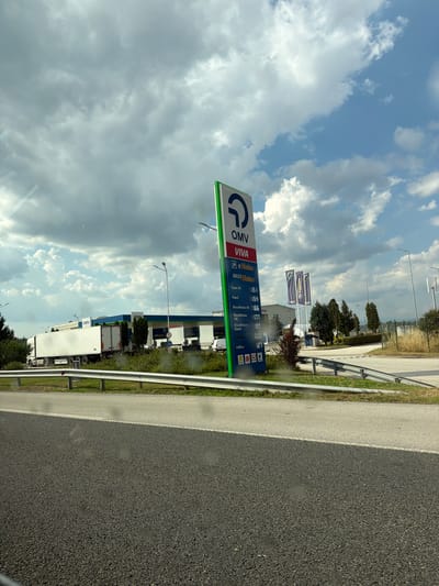The image captures a view of a highway near a gas station. In the foreground, a portion of the asphalt road is visible, marked by a white line and a metal guardrail. Beyond the guardrail, a grassy embankment leads to the gas station area.
The gas station itself features a modern design with a blue and white canopy. Several OMV logos are visible on the building and on flags. A tall, vertical sign displaying OMV and VIVA branding, along with fuel prices, stands prominently near the entrance. The prices listed include Super 95 at 1.54, Diesel at 1.66, MaxxMotion 95 at 1.77, MaxxMotion Diesel at 1.80, and AdBlue at 1.27. A white articulated lorry is parked to the left of the gas station. The sky above is partly cloudy with patches of blue. The location is identified as Shtarkovo, Bulgaria.