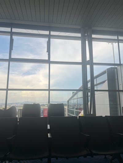 The image depicts an interior view of an airport departure lounge in Guangzhou City, China, as evidenced by the Chinese text on a poster visible on the wall. The scene is characterized by a large window offering a panoramic view of the airport tarmac and the sky. The window is divided into a grid by dark-colored framing, and through it, several airplanes can be seen on the ground and in the distance, along with the typical airport infrastructure such as control towers and jet bridges. The sky is partially cloudy with sunlight filtering through, suggesting it is daytime. In the foreground, several rows of dark-colored airport seating are visible, silhouetted against the bright exterior view. Some of these seats are occupied by unseen individuals, indicated by their presence. The overall atmosphere is one of waiting and transit, typical of an airport environment. The architecture suggests a modern airport terminal with extensive glass facades.