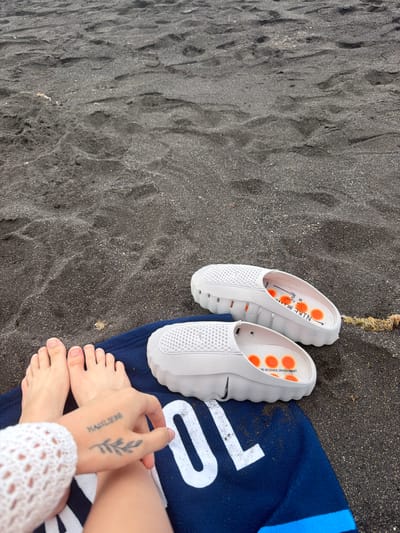 On a beach with dark, wet sand, a pair of white Nike slide sandals with orange accents sits on a dark blue towel. A person's feet, with a tattoo on their hand, are also visible on the towel. The scene is located in Puerto de la Cruz, Spain.