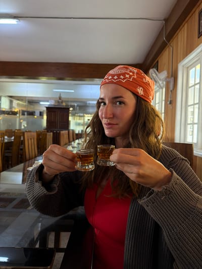 The image displays an adult female presenting two shot glasses within an indoor dining establishment located in Ramboda, Sri Lanka. The woman occupies the central foreground, characterized by long, wavy brown hair, blue eyes, and an orange bandana featuring a white "HARLEY-DAVIDSON CYCLES" pattern. She wears a red garment with a central zipper and a dark grey knitted cardigan. Her gaze is directed at the camera, exhibiting a slight smile. Each hand holds a small, clear glass shot glass containing an amber-colored liquid, with visible particulate matter or herbs. In the immediate foreground, a dark, reflective table surface is partially visible, with a smartphone resting on its left side. The background reveals an expanse of an empty dining area furnished with numerous wooden tables and high-backed wooden chairs. Behind this seating area, visible through a glass partition, is a dark wooden cabinet topped with a golden Buddha statue. The room's architecture includes a white ceiling with fluorescent tube lighting, a horizontal wooden beam, and a wood-paneled wall on the right side. This wall features multiple white-framed windows and a white wall-mounted oscillating fan. Exposed electrical conduit is visible on the ceiling and wall.