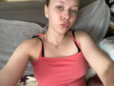 The media file is a close-up, rotated selfie of a young Caucasian woman lying on her back. She is wearing a pink tank top and a black bra. A delicate silver necklace with a clear pendant is visible on her chest. Her eyes are light blue, and she has a pouty expression. Her arms are stretched out to the sides, and one arm is partially obscured by her body. She appears to be indoors, possibly on a bed or couch, as indicated by the soft, textured blankets and the presence of a pillow. The lighting suggests it is daytime. There is no visible text in the image. The scene conveys a relaxed, casual mood.