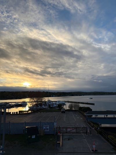 The image shows a view of a marina and lake at sunset or sunrise. The sun is obscured by clouds in the sky. In the foreground, there is a parking lot with parking spaces painted on it. Signs are visible, including one that says "Members Only, Laconia Yacht Club". A gate with a "Private Property - No Passing" sign is also present. Boats are docked in the marina along the shoreline. Across the lake, a tree-lined shoreline is visible. In the distance, a boat is traveling on the lake.