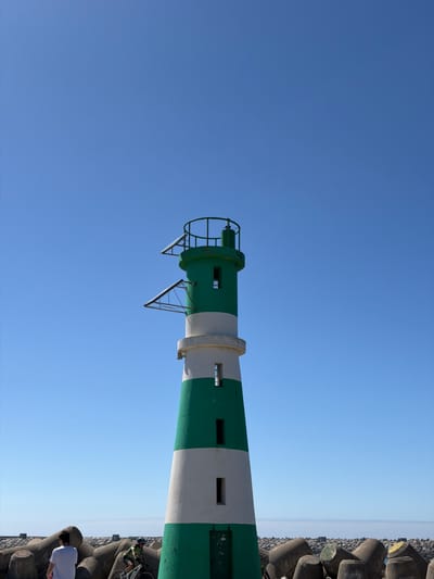 The image shows a lighthouse in Gafanha da Nazaré, Portugal, with a clear blue sky. The lighthouse is tall and cylindrical, painted with alternating bands of green and white. It has a white concrete base and a green metal railing at the top. There are two triangular solar panels attached to the lighthouse. In the foreground, there are concrete wave-breaking blocks. A person in a white t-shirt is walking away from the camera to the left. To the right, a person on a bicycle wearing a green cycling outfit and helmet is riding past the lighthouse. The background shows a hazy horizon with what appears to be the sea.