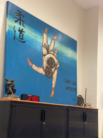 A painting depicting a judo scene hangs on a wall. A judoka throws their opponent onto a blue mat, with "Judo-Club Roenheim" and Japanese characters printed above and below. Next to the painting, a digital clock displays "21:02," and a small statuette sits below. A black cabinet with louvered doors stands to the right, and a decorative musical instrument rests on top of the cabinet.