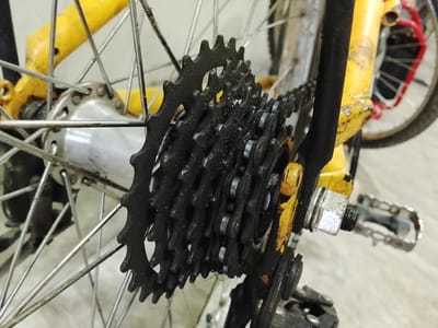 A close-up, slightly angled, eye-level shot shows the rear derailleur and cassette of a bicycle. The bicycle frame is predominantly yellow with some black and red accents visible on the rear wheel. The cassette consists of multiple black sprockets of varying sizes. A bicycle chain, also black, is visible engaging with the sprockets. The rear wheel spokes are silver. A metal nut and bolt assembly is visible securing a part of the bicycle frame. A portion of a silver pedal is also visible on the right side of the image. The background appears to be a light-colored, flat surface, possibly concrete or tile. This image was captured in Rumuepirikom, Nigeria.