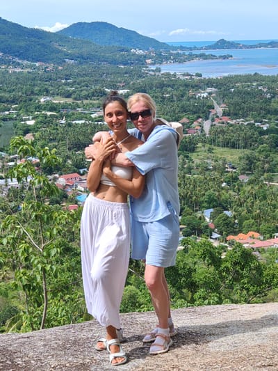 Two individuals are standing on a large rock formation, embracing. The person on the left, with dark hair pulled back, is wearing a white bandeau top and flowing white pants. The person on the right, with blonde hair and sunglasses, is wearing a light blue matching set of a loose-fitting top and shorts. They are both wearing sandals. Behind them, a panoramic view of a lush green landscape unfolds, characterized by dense palm trees and scattered buildings with orange and red tiled roofs. In the distance, a body of water, likely the sea, is visible, meeting the horizon under a bright, partly cloudy sky. This scenery is consistent with the location in Baan Harn, Thailand, known for its tropical terrain and coastal views.
