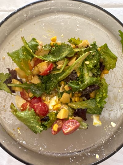 A nearly empty glass bowl of salad sits on a white tablecloth. The salad contains lettuce, corn, diced mango, halved cherry tomatoes, and chopped hard-boiled egg. The bowl shows evidence of prior consumption, with remnants and dressing residue visible on the interior surface. The location is presumed to be Celorico de Basto, Portugal. No people are visible.