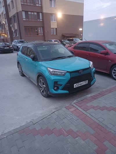 The image shows a teal Toyota Raize parked on a gray concrete surface next to a brick building. The car is facing right, with its front bumper and hood clearly visible. The license plate reads "X554HT 154". To the left of the Raize, a red car is partially visible. The background features a multi-story building with numerous windows and balconies. The ground is paved with gray and pink rectangular bricks. The lighting suggests it is either early morning or late evening, and the sky is overcast. The scene appears to be an urban or suburban parking area.