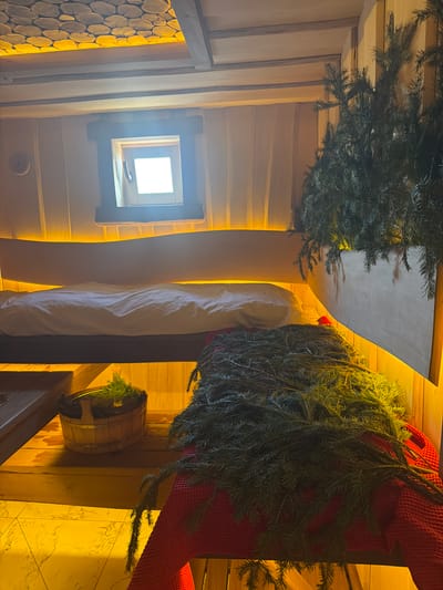 The image shows the interior of what appears to be a sauna, likely located in Марусино, Russia. The walls are constructed of light-colored wood planks. A small, square window is visible. A white cloth and a red textile are draped over what might be a bench or seating area. Green fir branches decorate the space, with one arrangement positioned above the window and another near the draped textiles. A small wooden bucket with greenery inside sits on a ledge or shelf. The lighting is warm and diffuse, illuminating the wooden surfaces.