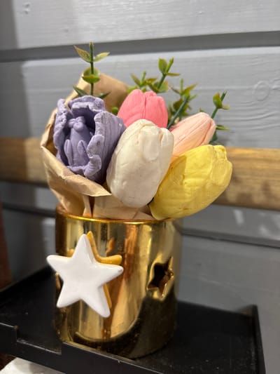 A close-up, eye-level shot showcases a decorative bouquet of artificial tulips. The tulips, made of a waxy material, are arranged in various pastel shades: pink, peach, yellow, white, and lavender. They are partially wrapped in a light brown paper. The bouquet is held within a gold-colored metallic object that appears to be a candle holder or lantern, with a star-shaped cutout visible on its surface. A white star decoration is attached to the front of the gold holder, with a smaller yellow star peeking out from behind it. The background features a light gray wooden surface, likely a wall or fence, with vertical planks. To the right, a dark object, possibly a lamp base or another decorative item, frames the scene. The lighting suggests an indoor setting during the daytime, with soft, diffused light. The image does not contain any visible text, and no people or specific activities are depicted, focusing solely on the still life arrangement.