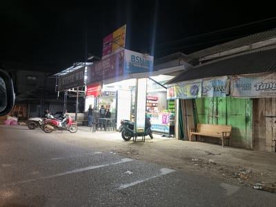 The image depicts a street scene in Keudepanteubreuh, Indonesia, at night. The street is lined with small shops or kiosks.

Several businesses are visible. "Kupie WD" and "Wanda Ponsel" are advertised. The signs indicate that Wanda Ponsel sells mobile phones and accessories, and that BSI Link is associated with Wanda Ponsel. One sign mentions assistance with biometric registration.

Several people are visible in front of the left-most kiosk. There are also several motorbikes parked. One black motorbike is parked in front of a store with a sign that says "ANDA PONSEL." Further down, there's a wooden bench.

The street is paved but appears weathered. There is some debris and litter on the ground. Streetlights illuminate the scene.