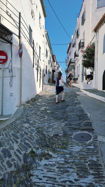 The image depicts a narrow, cobblestone street in Cadaqués, Spain, with whitewashed buildings on either side. The street slopes upwards, and there are people walking along it. In the foreground, a woman wearing a dark dress and a hat walks away from the camera, carrying a canvas bag. Further up the street, another woman in a hat and shorts walks towards the camera, while a man in a white shirt and shorts walks up a set of white stairs on the right side of the street. The buildings are traditional Mediterranean style, with balconies and windows. The sky is a clear blue, and there is a single tree with pink flowers on the right side of the street. The cobblestones are uneven and weathered, and there is some greenery growing between them. A manhole cover is visible in the cobblestone pavement on the right side of the street. There are electrical wires strung across the street overhead.
