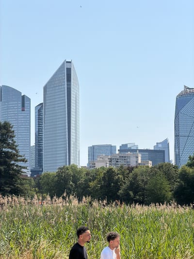 The image shows a view of the La Défense business district in Nanterre, France, on a clear, sunny day. In the foreground, tall reeds and green grasses line a path where two young men are walking. The man on the left wears a black t-shirt and is seen in profile, looking to the right. The man on the right wears a white t-shirt and is also seen in profile, with his hand on his chin. Behind the reeds and men, a dense line of green trees forms a backdrop. Beyond the trees, several modern skyscrapers of varying heights and designs dominate the skyline, characteristic of the La Défense district. The sky is a pale blue with two birds visible in flight.