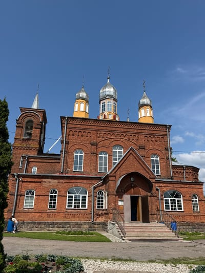 The image shows a brick Orthodox church under a clear blue sky with scattered clouds. The church has multiple onion domes, some silver and some gold and silver, topped with crosses. The facade is red brick with arched windows. A bell tower is visible on the left side of the building. There is a set of stairs leading up to the entrance, with railings. A person is visible on the left side of the building, near a large blue barrel, and appears to be bending over. Green trees and grass surround the church, with a gravel path in the foreground. The image is taken from a low angle, looking up at the church. The scene is likely the Church of the Assumption of the Blessed Virgin Mary in Chudovo, Russia, based on the architectural style and potential location.