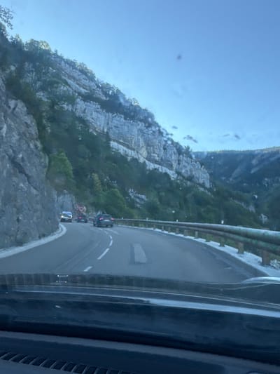 A car is driving on a winding road in a mountainous area. The road is paved and has white lane markings. On the left side of the road, there is a steep cliff face covered in rocks. On the right side of the road, there is a guardrail. The area is covered in trees and vegetation. The sky is a clear blue. The photo was taken from inside a vehicle, looking out through the windshield. The location is Septmoncel les Molunes, France.