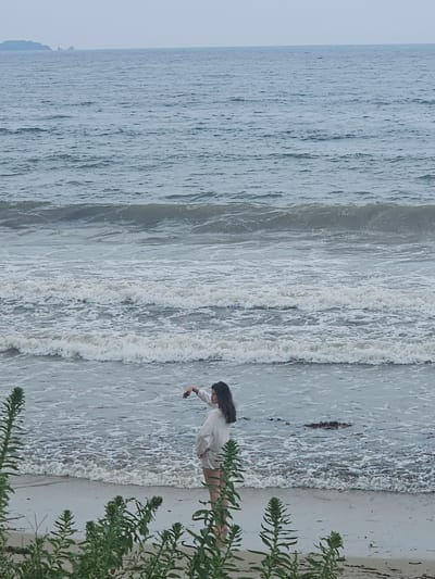 A woman stands on a sandy beach in Fokino, Russia, facing the sea. She is wearing a light-colored shirt and shorts. Her left arm is raised, and she appears to be holding a phone, possibly taking a selfie or a photo. The ocean has moderate waves with whitecaps, and the water is a muted blue-gray color. In the distance, a hazy landmass or island can be seen on the horizon. Green vegetation with tall stalks is in the foreground, partially obscuring the beach and the woman. The sky is overcast, contributing to the muted tones of the scene. The overall impression is one of a serene, slightly moody coastal day.