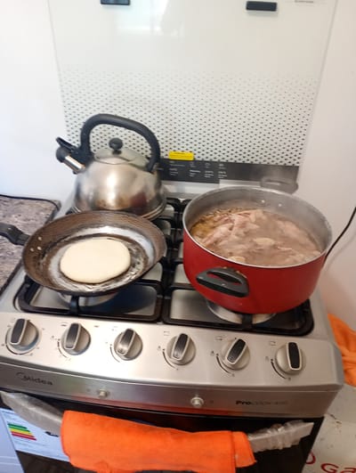 The image displays an indoor kitchen scene centered around a silver Midea ProCOOK-400 gas stove, where a meal is being actively prepared. The stove features four burners, three of which are currently in use.
On the left front burner, a dark frying pan contains a single, round, light-colored flatbread, likely an arepa, cooking on its surface. To the right, a shiny stainless steel kettle rests on the back left burner. On the right front burner, a large, red pot is actively boiling, with visible steam rising from its contents. Inside the pot, pieces of meat, possibly chicken or pork, are simmering in a broth, accompanied by what appear to be pieces of onion or garlic.
The stove has multiple control knobs with clear markings for flame adjustment. Below the stove, an orange microfiber cloth is draped over part of the oven door or a lower drawer. To the left, a portion of an energy efficiency label is visible, showing the word "ENERGIA" and a series of color-coded bars.
The background consists of a clean white wall or backsplash adorned with a repeating pattern of small, dark triangles. Above the stove, a panel on the backsplash displays the "Midea" brand name and various cooking instructions or oven settings, including temperature indications such as "170°C", "210°C", and "190°C". The scene conveys a functional and lived-in kitchen environment, indicative of a home in Pozo Almonte, Chile.