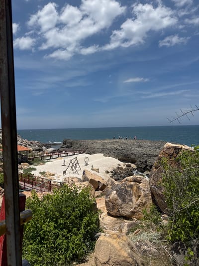 This image depicts a scenic coastal area in Vinh Hai, Vietnam. The foreground is partially obscured by a rustic wooden structure with a red and yellow railing, and lush green foliage. Beyond this, a white pebble beach meets a rugged, rocky shoreline. A wooden walkway with red railings traverses the beach and connects to a small gazebo with an orange roof. On the beach, a swing set and two heart-shaped sculptures are visible. In the middle ground, a group of people are standing on a rocky promontory overlooking the ocean. The sea is a deep blue, with moderate waves, and a clear sky with scattered white clouds stretches across the horizon. The overall scene conveys a tranquil, natural environment suitable for recreation and sightseeing.