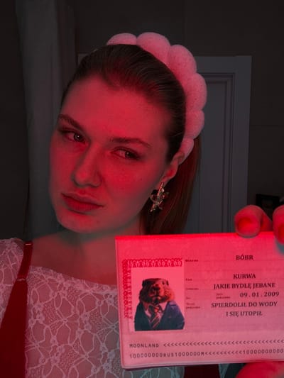 A person illuminated by a red light holds up a Polish ID-like document. The document contains phrases in Polish, including "BÓBR," "KURWA," and "JAKIE BYDLĘ JEBANE." It has an official format with a photo of a man in a suit, a date, and text that translates to "drowned in water." The bottom includes the word "MOONLAND" and a sequence of "<<" and numbers followed by "RUS" and "M". The person holding the document is wearing a headband and a white lace top.