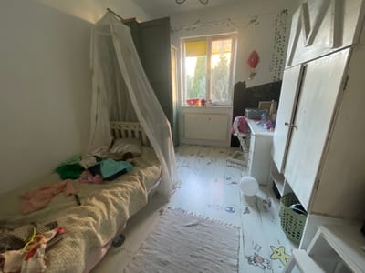 This is a wide-angle indoor shot of a child's bedroom in Belarus. The room is brightly lit by natural light coming from a large window. The walls are white, and the floor is covered with decorative stickers of animals and words like "moon," "elephant," and "ball."

The room features a crib covered with a mosquito net, a bed with a textured blanket and various clothes strewn across it, and a large white wardrobe with an "M" shape on its doors. A small desk with a chair, a toy hot air balloon hanging on the wall, and a green laundry basket are also visible. A white rug is placed on the floor.

The overall impression is of a well-used, lived-in child's room, likely during the daytime given the bright natural light. The presence of toys, clothes, and the crib suggests it is a functional living space for a child. The room appears to be in the process of being tidied or used, with items out of place.
