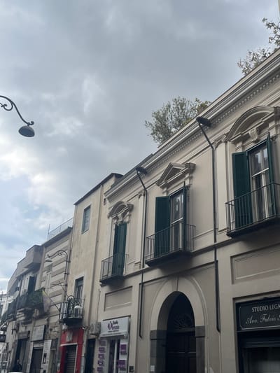 An urban street scene in Scafati, Italy, depicts a row of multi-story buildings under an overcast sky. The prominent structure on the right is a cream-colored building with neoclassical architectural features, including ornate cornices, decorative window surrounds, and multiple windows fitted with dark green louvered shutters and wrought-iron balconies. A large, dark arched entrance is centered on its ground floor. To the right of this arch, a sign reads "STUDIO LEGALE Avv. Fulvio S."; to the left, a sign for "Studio Virtusio - Centro Odontoiatrico" is displayed. Two black, modern streetlights are mounted on the building's facade. Green vegetation, likely trees or shrubs, is visible along its flat rooftop.

To the left, additional buildings extend down the street. One adjacent building features a striped facade pattern and a balcony. Further down, another building has a balcony from which a vertical flag or banner, colored yellow and purple, is suspended.

In the upper-left quadrant of the frame, a traditional black street lamp with a curved arm and a single downward-facing light fixture is visible. At the lower-left corner, a person dressed in dark attire is partially visible, walking on the sidewalk.