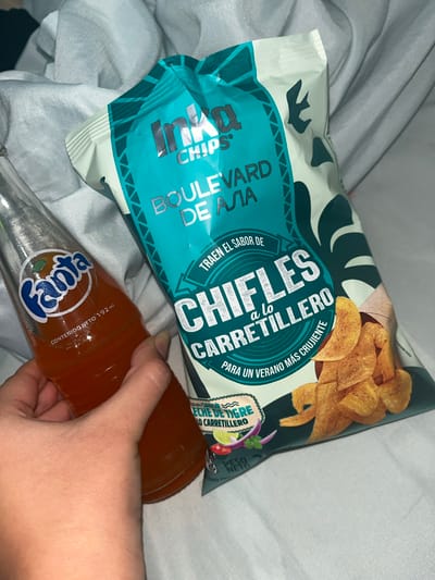 A hand holds a glass bottle of Fanta while resting on a white, wrinkled cloth. A bag of Inca Chips is positioned next to the bottle. The bag is teal and white with text and images of plant leaves and a bird. The Fanta bottle is orange and has a white and blue label. The scene appears to be indoors, possibly a bedroom or a studio.