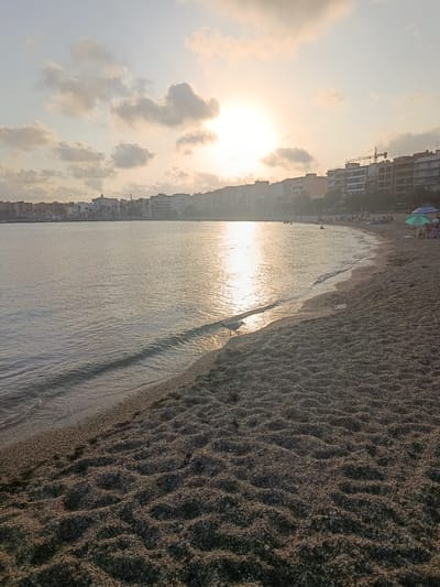 The image depicts a coastal scene at sunset in Águilas, Spain, likely along the Playa de Levante or a similar urban beach. The sun is setting behind scattered clouds, casting a warm glow on the water and the sky. Buildings line the distant coastline, with some under construction, indicated by a visible crane. The beach is composed of dark pebbles and sand, with footprints visible in the foreground. Gentle waves lap at the shore. A single seagull stands near the water's edge. A few beach umbrellas and people can be seen further down the beach, suggesting a relaxed atmosphere. The overall lighting suggests late afternoon or early evening.