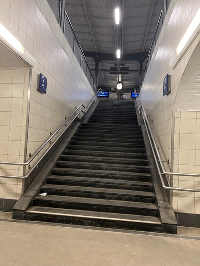 This is a vertical, eye-level shot looking down a long set of concrete stairs in what appears to be a public transportation station in Verden (Aller), Germany. The stairs are flanked by metal handrails and are bordered by tiled walls and floors. Above, the ceiling is corrugated metal, and fluorescent lights illuminate the scene.
On the tiled walls, there are blue signs with white numbers: a "2" is visible on the upper left, and a "3" is visible on the lower left. A clock is also visible on the wall near the middle of the stairs, suggesting it is daytime. A few digital screens are also present. The overall impression is of a functional, utilitarian public space. There are no people visible in the image, and the scene is clean but shows some signs of wear on the stairs. The weather is not discernible.