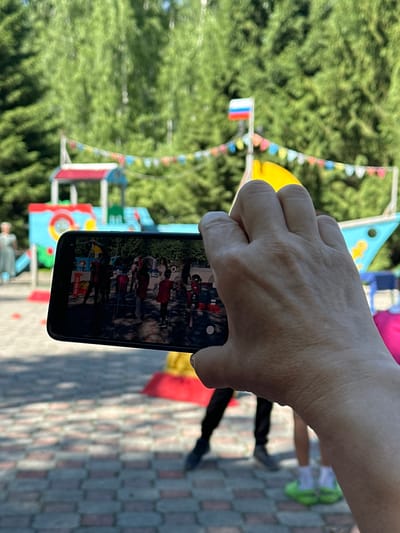 A person's hand holds a smartphone, capturing a video of a group of children gathered in an outdoor play area. The children are in the center of the frame on the phone's screen, some appearing to be wearing red shirts. In the background, behind the phone, is a colorful playground structure resembling a ship, decorated with a string of festive flags and a Russian flag flying from a pole. The ground is paved with brick-like tiles. The environment appears to be a park or recreational area with lush green trees forming a backdrop. The overall scene suggests a daytime event or gathering in a public outdoor space.