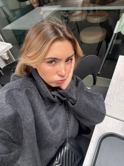 A young adult female is depicted in a medium-close-up selfie, seated at a light-gray table within an indoor commercial establishment, likely a cafe or restaurant, located in Vaulx-en-Velin, France. The subject has blonde hair with darker roots, brown eyes, and is wearing a dark charcoal-gray, textured, hooded garment. Her right hand (viewer's left) rests against her cheek, and her lips are pursed in a pout, directed towards the camera. A black quilted handbag with a silver-colored chain strap is visible across her body. On the table to her right (viewer's left), a black serving tray is present. The background consists of reflections in a glass panel, revealing elements of an interior space, including a young child with light hair looking away from the camera, two round light-colored stools, and a light-colored high chair.