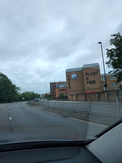 This image depicts an urban street scene in Peterborough, United Kingdom, specifically showing the UNEX Crescent College building located near Crescent Bridge. The perspective is from inside a vehicle, with the top of a dashboard visible at the bottom of the frame.
The sky above is uniformly overcast, exhibiting a light grey color. An asphalt road, marked with white dashed lane lines and white directional arrows, curves gently to the left from the foreground into the distance. A metal barrier fence separates the road from the sidewalk and buildings on the right.
On the left side of the road, a dense line of mature green trees borders the thoroughfare, partially obscuring the distant background. Two traffic light poles with active green signals are visible amidst the foliage on the left.
On the right side of the road, a prominent multi-story brick building with a grey roof section dominates the midground. Vertically aligned large black letters spell "UNEX" on its left-facing facade. Below "UNEX," a logo featuring a crescent moon and a lion accompanies the text "CRESCENT COLLEGE." Further to the right on the same building, large black letters spell "PLANT A TREE" vertically. The building features various rectangular and square windows, some with visible white frames, and several external air conditioning units near its base. A tall, black street lamp pole stands directly in front of this building on the right side. Behind this primary structure, additional brick buildings are visible.
In the distance, on the road ahead, a light-colored vehicle and a darker vehicle are discernible. No individuals are visible in the scene.