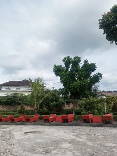 Red concrete benches line a paved area facing a low hedge. Behind the hedge are various trees and shrubs, including a palm tree. A light-colored building with a dark roof is visible in the background on the left side. Above the trees, a cloudy sky fills the frame.